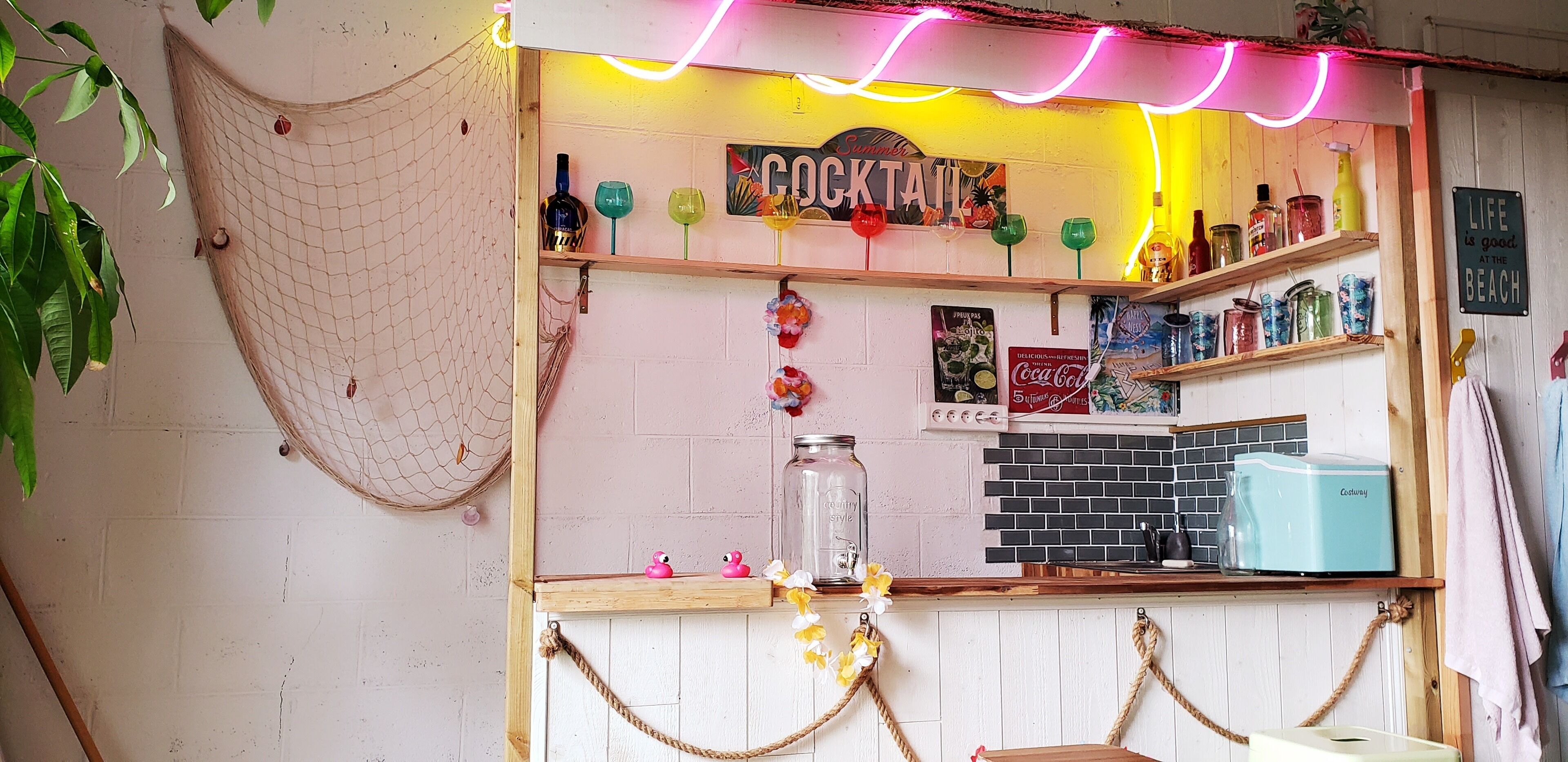 A cocktail bar with a neon sign, shelves with bottles, and a Coca-Cola cooler.