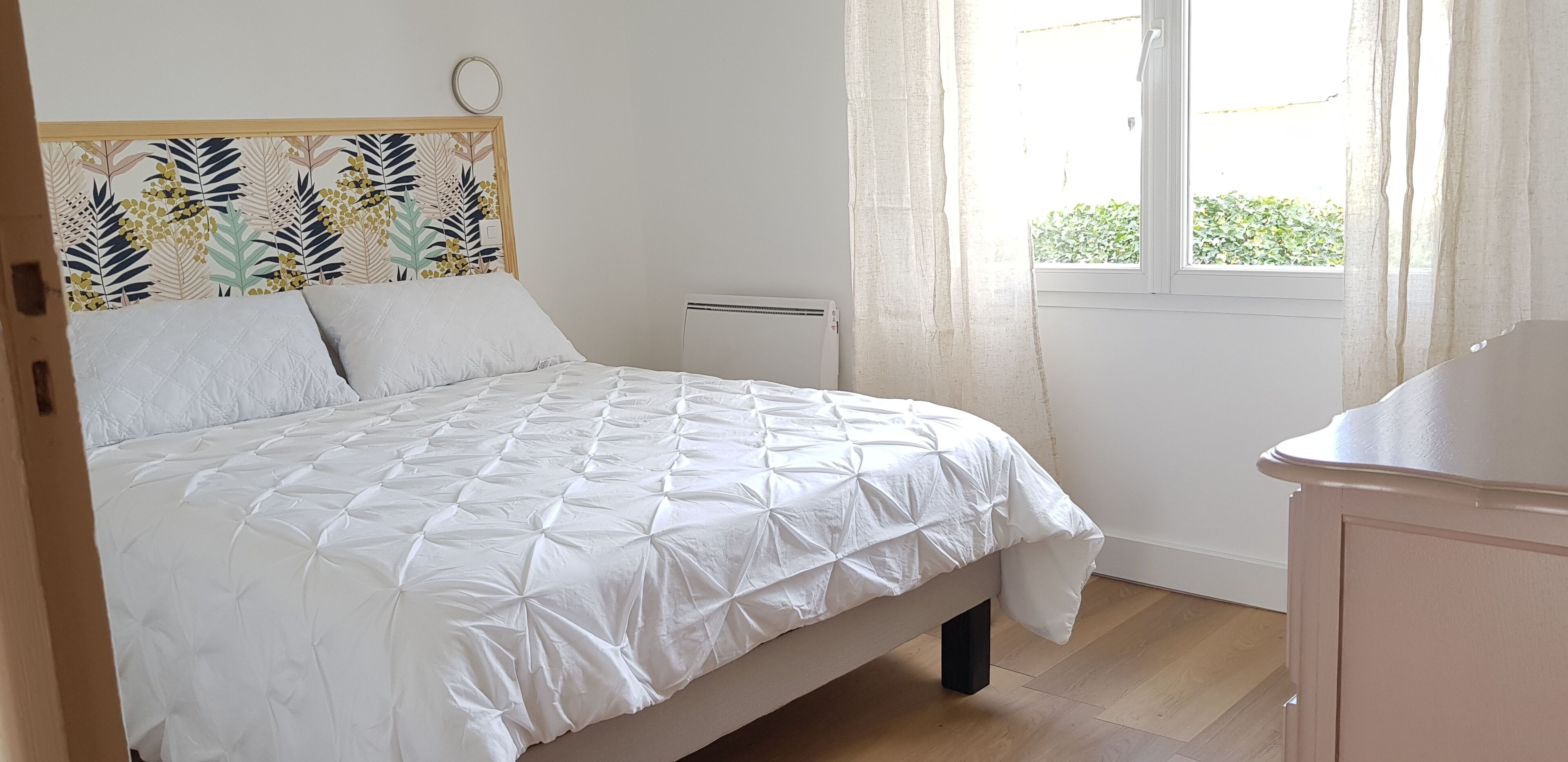 Een netjes opgemaakt bed met een witte quilt, een hoofdbord met een bladmotief en een raam met gordijnen.