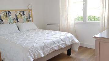 Een netjes opgemaakt bed met een witte quilt, een hoofdbord met een bladmotief en een raam met gordijnen.