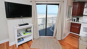 Una sala de estar con televisor de pantalla plana, un mueble blanco para equipo audiovisual con libros, un tapete estampado y vista a la playa a través de un ventanal corredero.