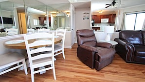 Una sala de estar con un sillón reclinable de cuero marrón, una mesa de comedor blanca con sillas y una cocina al fondo.