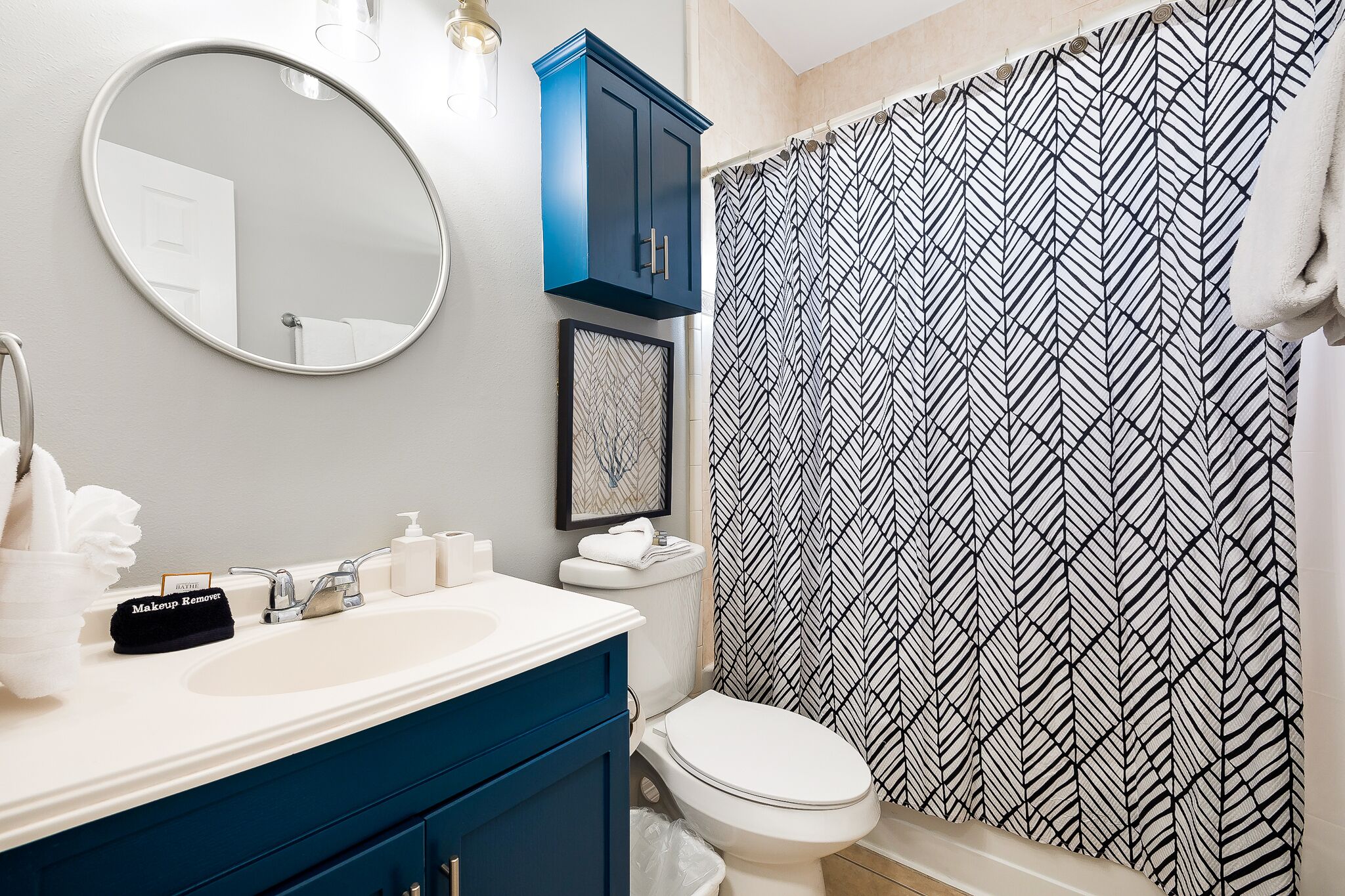 A bathroom with a white sink, a blue cabinet, a toilet, a mirror, and a black and white patterned shower curtain.