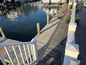 A wooden dock extending into calm water, with a white railing and potted plants along the side.