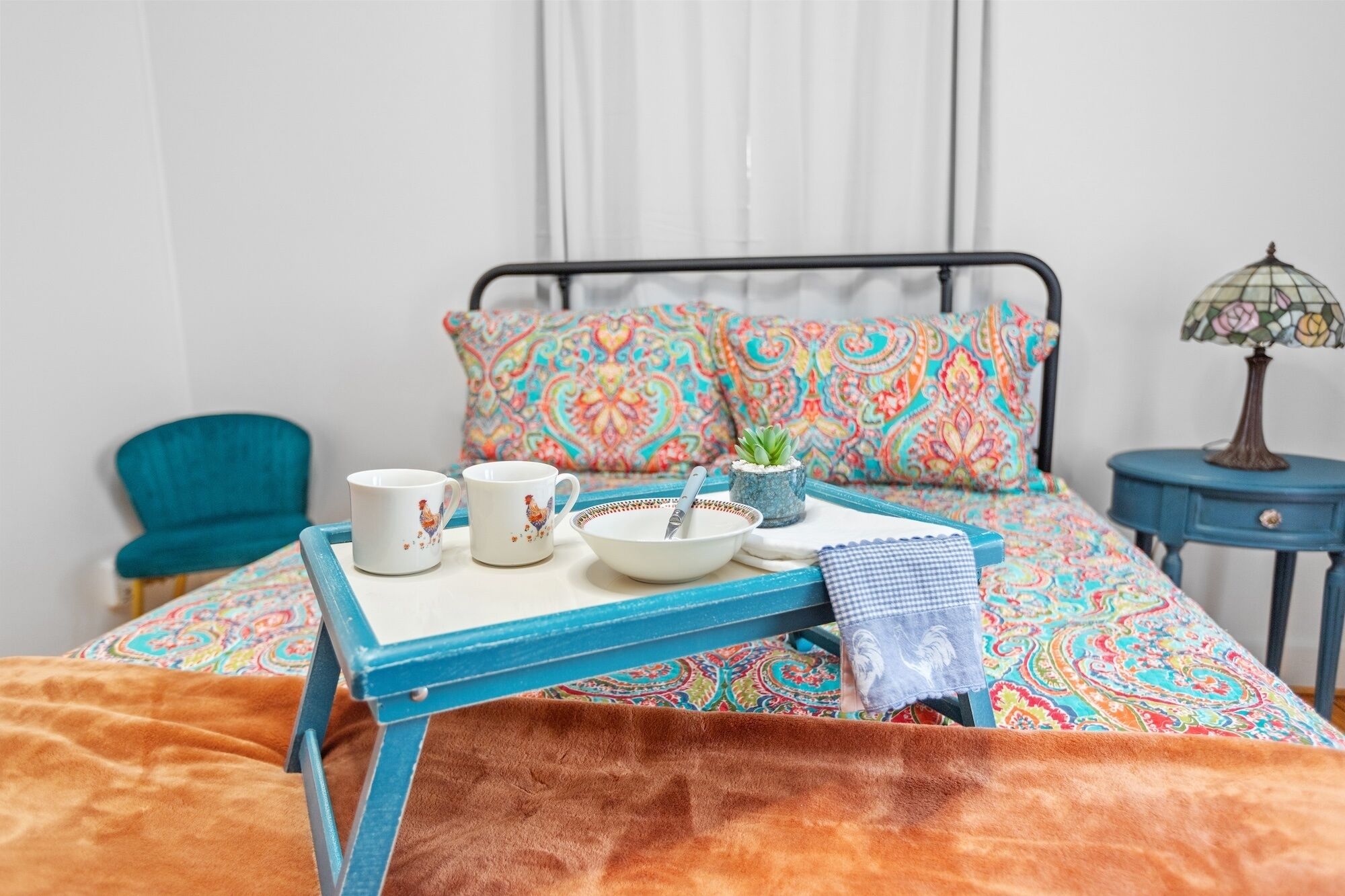 A neatly made bed with a colorful patterned bedspread, a blue metal tray with a bowl and two white mugs, a small potted plant, and a lamp on a side table.