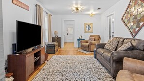 A living room with a flat-screen TV, a wooden cabinet, a blue side table, a brown leather sofa, and a patterned rug. - Deannies Cottage 2 Bedroom Cottage by RedAwning (Fredericksburg)