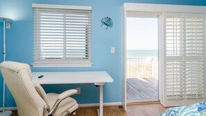 Quarto com escrivaninha, cadeira, janela com persianas e porta que dá para uma varanda com vista para a praia.