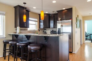 A modern kitchen with dark cabinetry, a central island with stools, a stainless steel refrigerator, and pendant lighting.
