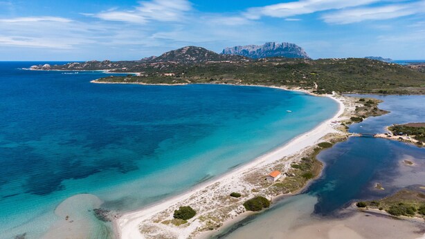 An aerial view of a coastal area with a beach, clear blue waters, and a mountain in the background.