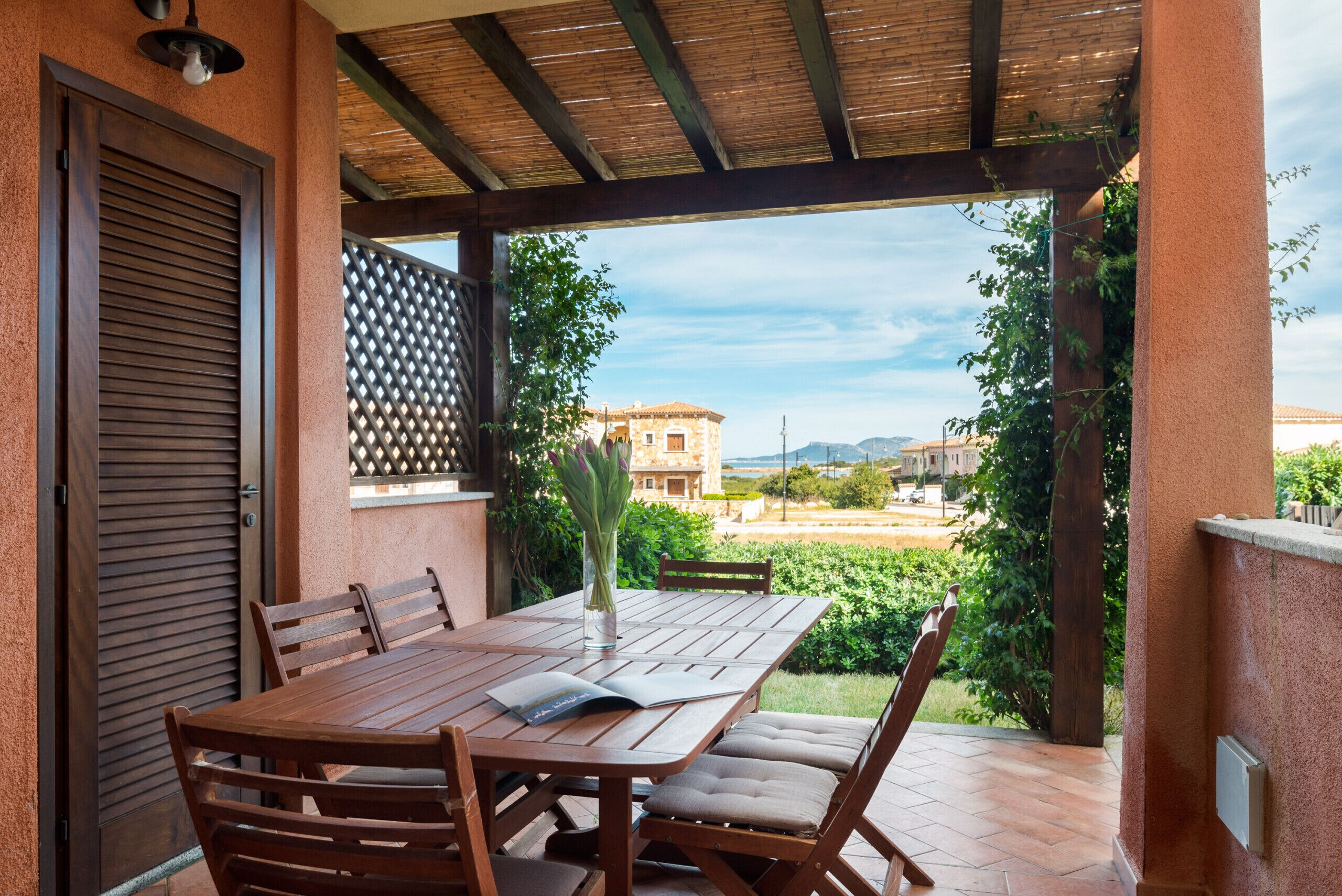 A wooden table with chairs and a vase of flowers, set on a patio with a view of buildings and greenery.