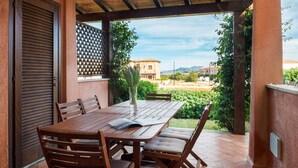 A wooden table with chairs and a vase of flowers, set on a patio with a view of buildings and greenery.