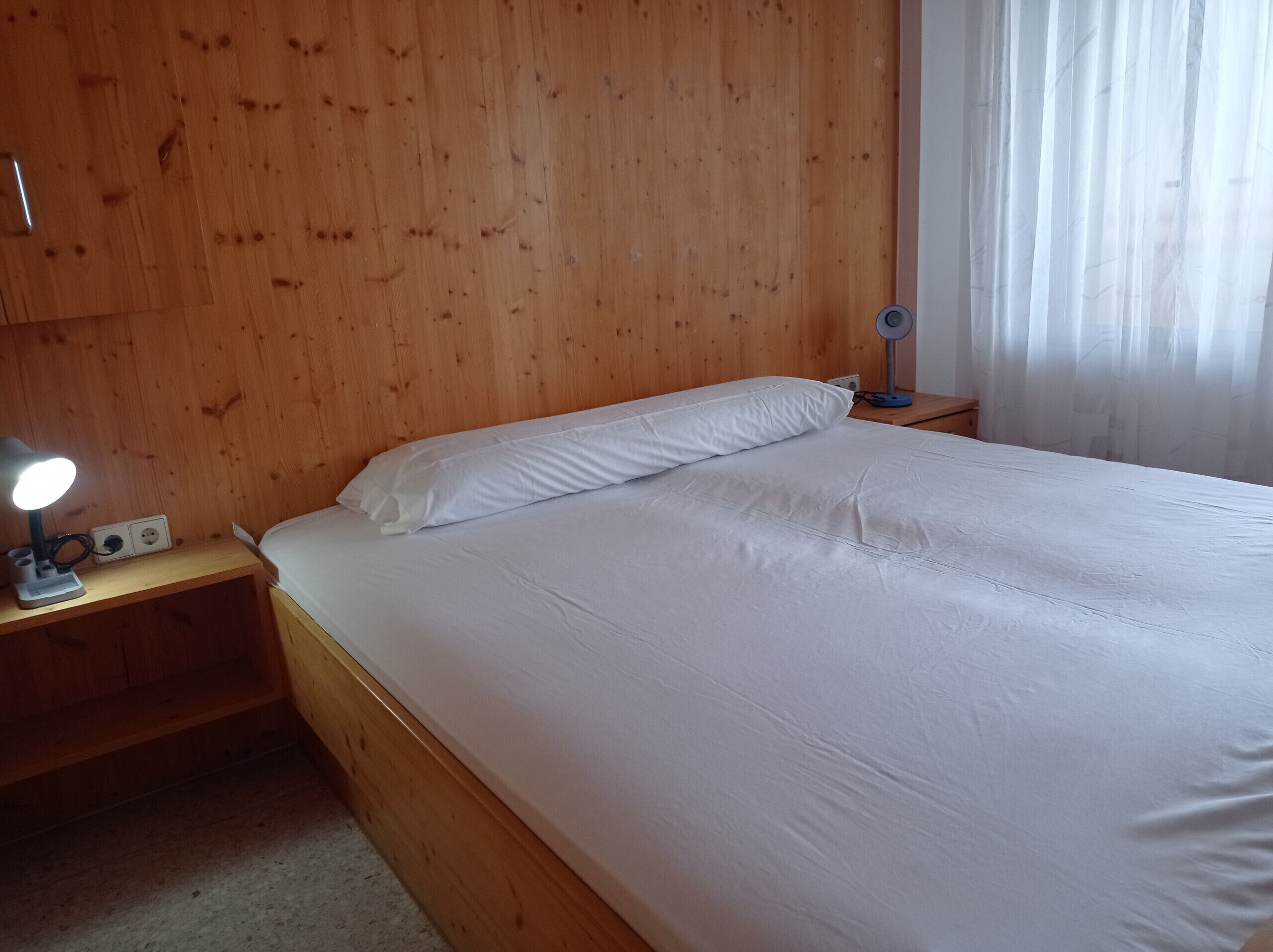 A wooden bed with white bedding, a bedside lamp, and a power outlet on a wooden nightstand.