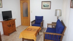 Een woonkamer met een televisie op een houten kast, een bank met blauwe kussens, een houten tafel met een gele tafelkleed en een lamp op een bijzettafeltje.