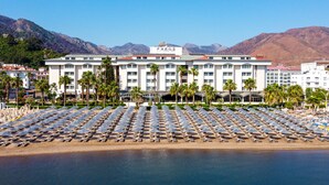 Un hotel con una amplia zona de playa que cuenta con filas de tumbonas y sombrillas, un edificio destacado con la señalización 'Faros Premium Beach' y un paisaje montañoso de fondo.