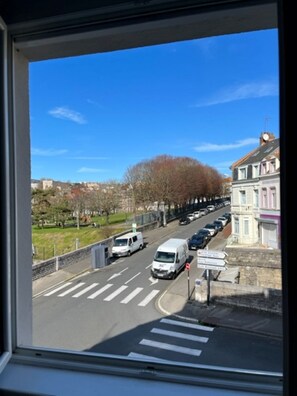 A street view from a window with a clear blue sky, a pedestrian crossing, parked cars, and buildings in the background.