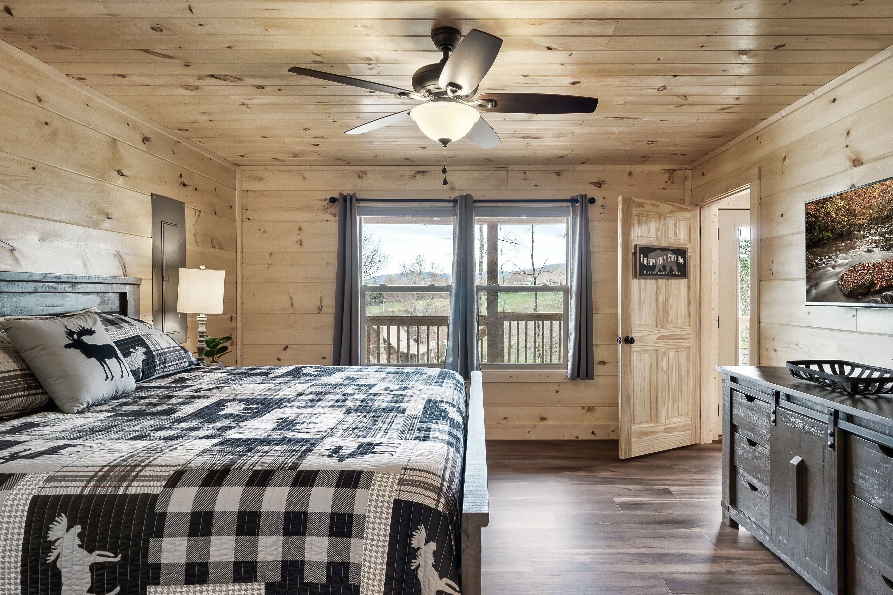 Un dormitorio de cabaña de madera con una cama, ventilador de techo, ventana con cortinas, una puerta y vistas a una terraza.