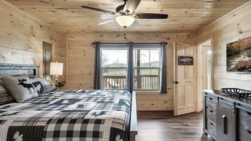 Un dormitorio de cabaña de madera con una cama, ventilador de techo, ventana con cortinas, una puerta y vistas a una terraza.
