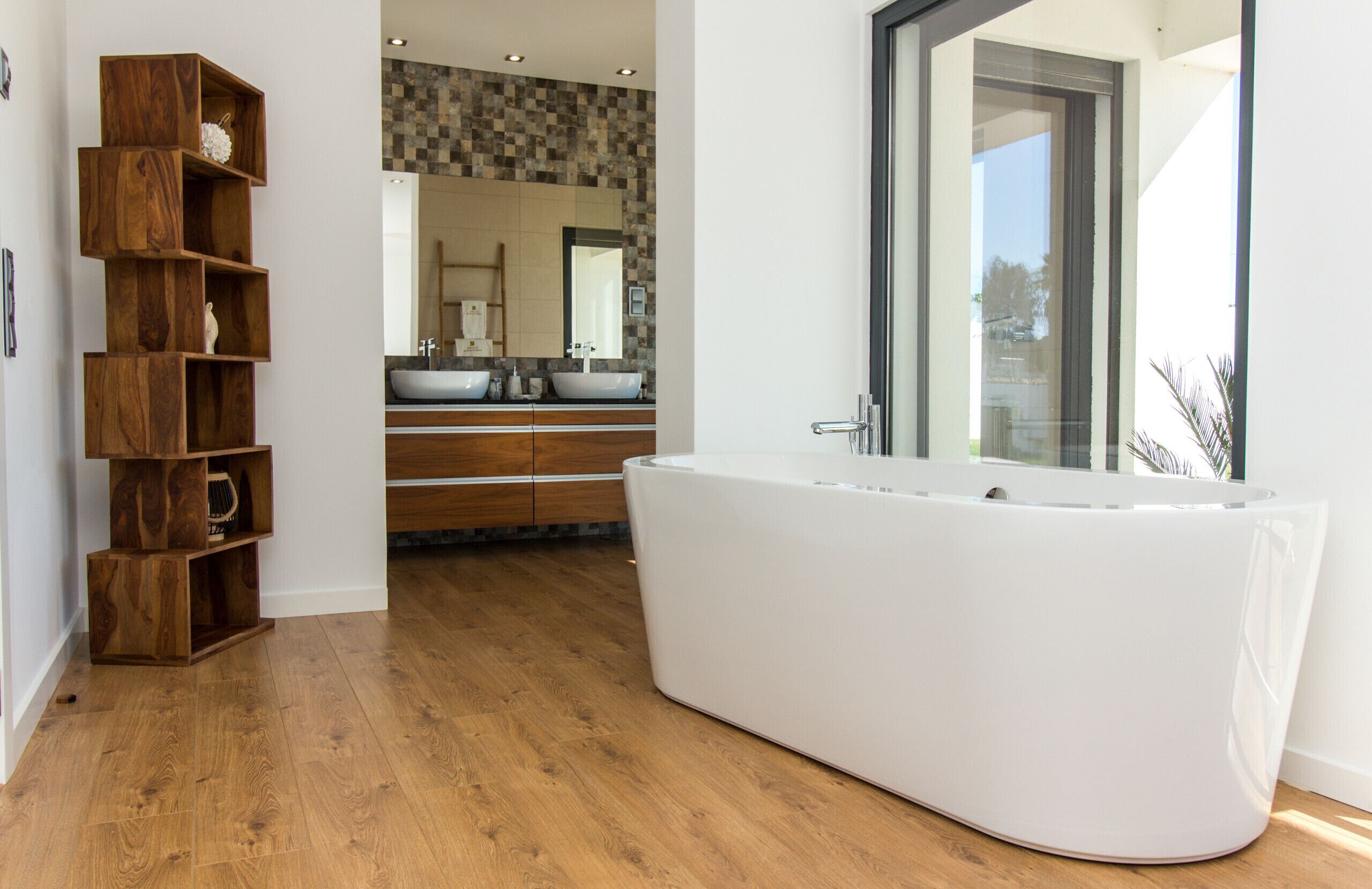 Un baño moderno con una bañera grande, estantes de madera, un tocador con lavamanos y un espejo amplio.