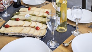 Un coin repas avec une table dressée pour un repas, comprenant une variété de fromages et de cornichons, deux bouteilles de vin et plusieurs verres.