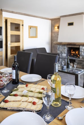 Uma sala de jantar com mesa posta para refeição, exibindo diversos queijos, picles, duas garrafas de vinho e vários copos.