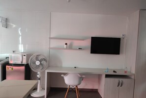 Una cucina compatta con forno a microonde, frigorifero e una ventola bianca. C'è una scrivania bianca con una sedia e una TV montata a parete.