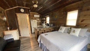 Una stanza in un cottage di legno con un letto, una scrivania, una sedia, angolo cottura e una finestra con persiane.