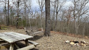 Un tavolo da picnic rustico e un'altalena di legno in una zona boschiva, con un braciere nelle vicinanze.