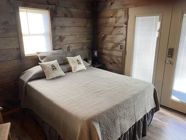 A bedroom with a wooden log wall, a bed with a textured blanket, two pillows with bee designs, a nightstand with a lamp, and a door with a window.