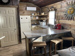 A kitchen with a granite countertop, wooden stools, a refrigerator, a microwave, a toaster, and a wooden wall with a sun decoration.