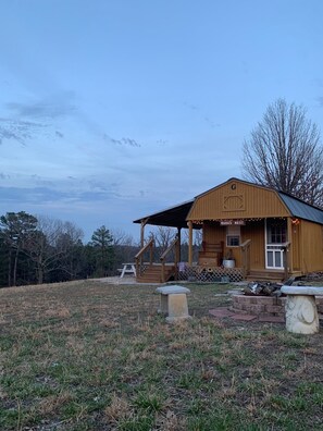 A wooden cabin with a porch, a sign reading "Manas Nest," and a picnic table in a grassy field.