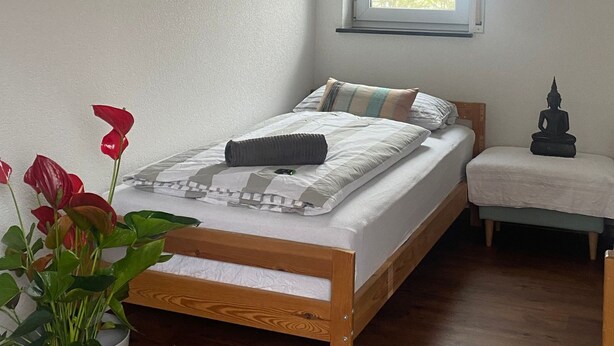 Quarto com cama de madeira, janela, um pequeno banco e um vaso com planta.