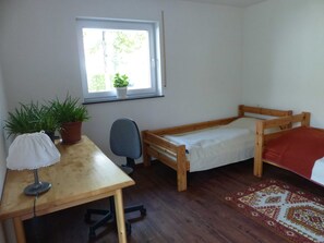 Ein kleiner, ordentlicher Raum mit einem Holzbett, einem Schreibtisch mit Stuhl, einer Topfpflanze und einem Fenster mit Blick auf Bäume.