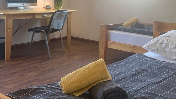 A bedroom with a wooden bed, a desk with a computer, a chair, a window, and a wall-mounted TV.