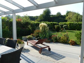 Un patio al aire libre con una mesa y sillas de madera, rodeado de plantas en macetas y vistas a un jardín.