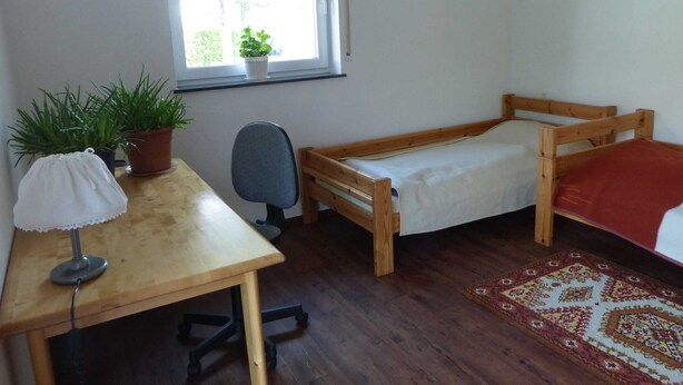 Una habitación pequeña y ordenada con una cama de madera, un escritorio con una silla, una planta en maceta y una ventana con vistas a árboles.