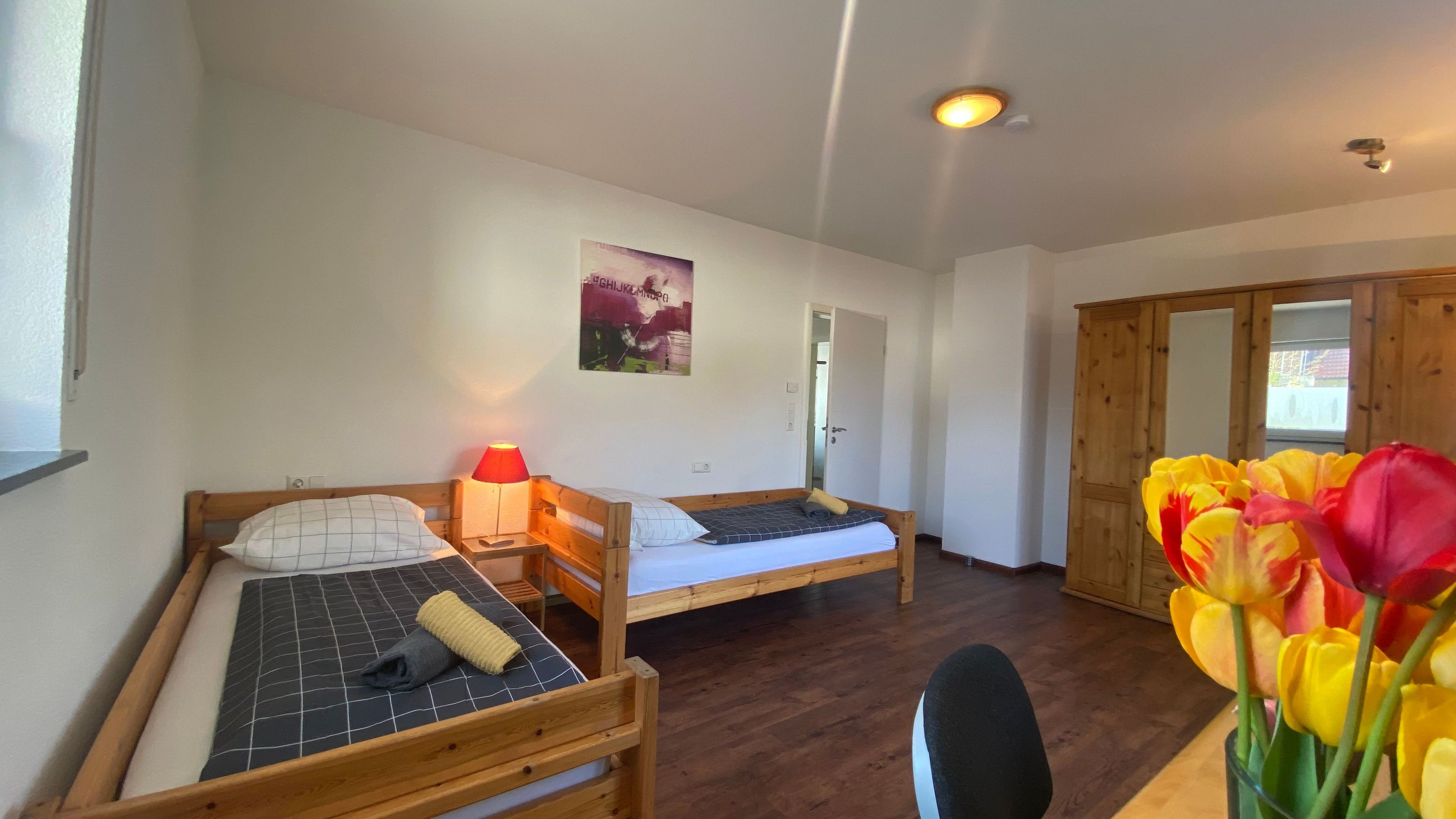 A room with two wooden beds, a small table, a chair, a wall-mounted lamp, and a door leading to another room.
