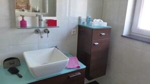 Une salle de bain avec un lavabo blanc, un miroir, une étagère en verre où sont rangés les articles de toilette et un meuble en bois foncé.