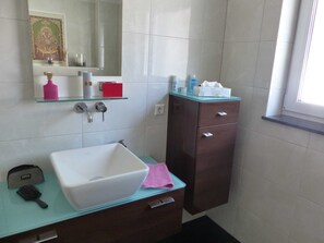 Ein Badezimmer mit einem weißen Waschbecken, einem Spiegel, einer Glasablage für Toilettenartikel und einem dunklen Holzschrank.