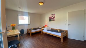 Een knus kamertje met twee houten bedden, een bureau met stoel, een raam en een deur.
