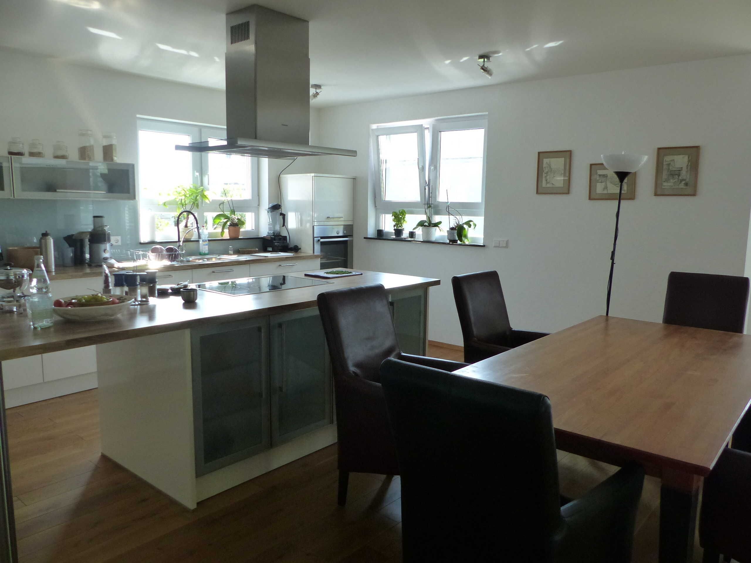 Une cuisine moderne dotée d’un îlot central, d’un coin repas avec une table et des chaises en bois, et offrant une vue sur l’extérieur grâce à de grandes fenêtres.
