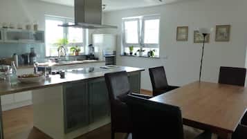 Eine moderne Küche mit einer zentralen Kochinsel, einem Essbereich mit Holztisch und Stühlen und Blick ins Freie durch große Fenster.
