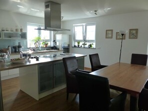 Une cuisine moderne dotée d’un îlot central, d’un coin repas avec une table et des chaises en bois, et offrant une vue sur l’extérieur grâce à de grandes fenêtres.