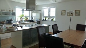 A modern kitchen with a central island, a dining area with a wooden table and chairs, and a view of the outdoors through large windows.