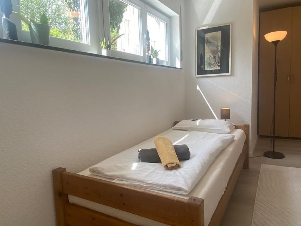 Un lit simple avec une tête de lit en bois, une table de chevet, une lampe sur pied et une fenêtre ornée de plantes.