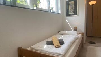 Un lit simple avec une tête de lit en bois, une table de chevet, une lampe sur pied et une fenêtre ornée de plantes.