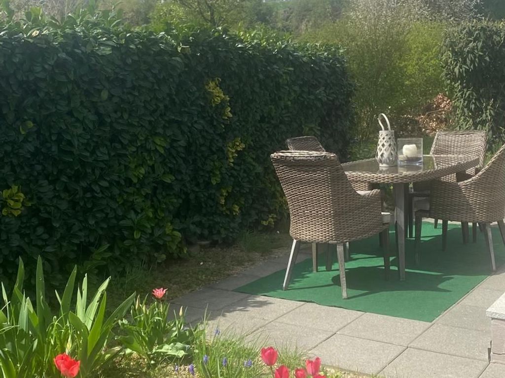 Une terrasse extérieure avec des chaises et une table en osier, entourée d’une haie et d’un jardin.