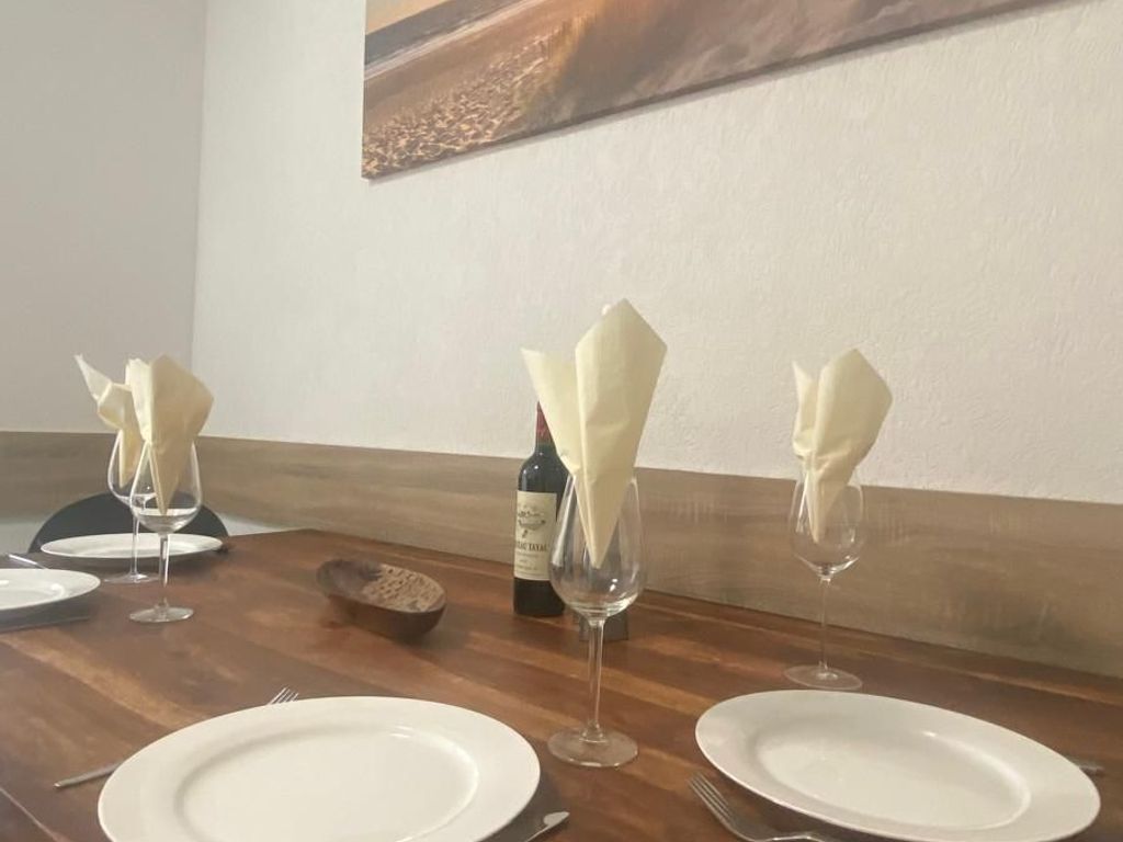 Una mesa de comedor dispuesta con platos blancos, copas de vino y servilletas dobladas. Se observa una botella de vino sobre la mesa.