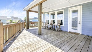 A covered deck with wooden railings, a seating area with chairs, and a door with glass panels. - The Neon Moon - 3rd Row Coastal beauty, close to the beach w/ Gulf Views! (Port Bolivar)