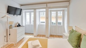 A modern bedroom with a bed, a dresser, a TV, and a view of the outside through large windows. - Bonjardim Studio Balcony by DA'HOME (Porto)