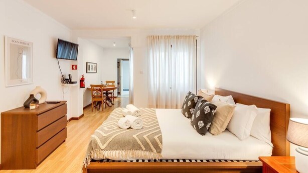 A bedroom with a bed, a wooden dresser, a TV, a desk, and a chair.
