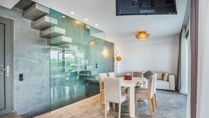 Un interior moderno con una escalera de cristal, un comedor con mesa y sillas blancas, y un sofá al fondo.